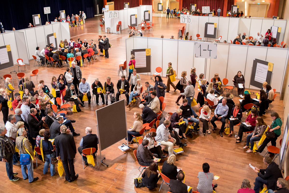 Open-Space-Veranstaltung mit Teilnehmenden, orangefarbenen Stühlen und Themen-Stellwänden in einer Halle